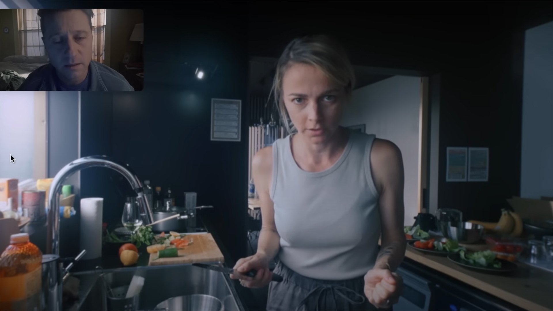 A woman seen on a video call stands in her kitchen holding a knife. In the top left is an image of her husband on the other end of the call.