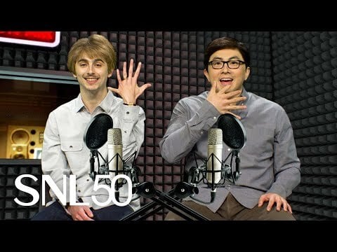 a white man with brown hair holding up a hand with six fingers. Next to him is an asian man in glasses wearing a button down shirt. Both are in a recording studio and surrounded by podcast equipment.